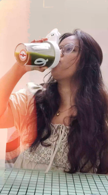 A woman with long dark hair and glasses drinking from a white plastic travel tumbler with a flip-top lid, set against a softly blurred indoor background.