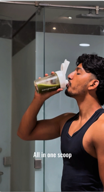 A man in a black tank top drinking from a clear shaker bottle with a white flip-top lid in a bathroom setting. The bottle has a green label with the brand name 'evergreen'.