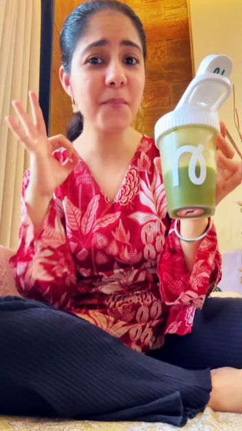 A woman sitting indoors holds up a green reusable tumbler with a white lid and a white logo, while wearing a red floral top and black pants.