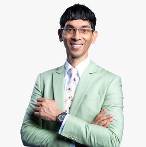 A man wearing a light sage green suit jacket, white dress shirt, and a white tie with a red and blue floral pattern, standing with arms crossed against a plain white background.