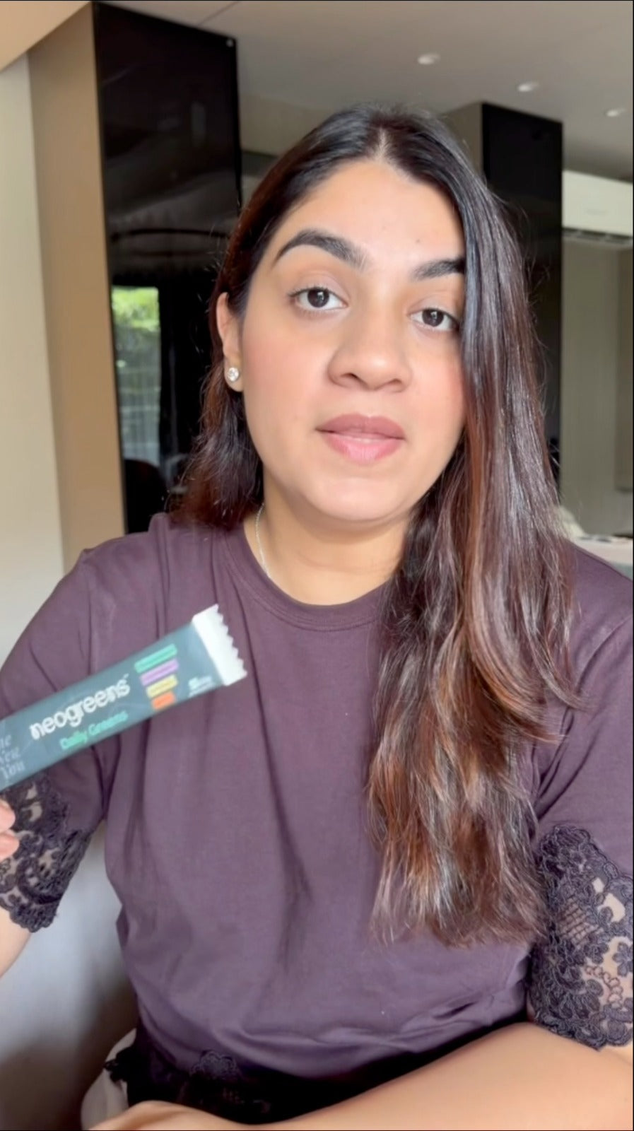 A woman with long brown hair wearing a purple t-shirt with lace sleeves, holding a Neogreens dietary supplement packet in a home setting.