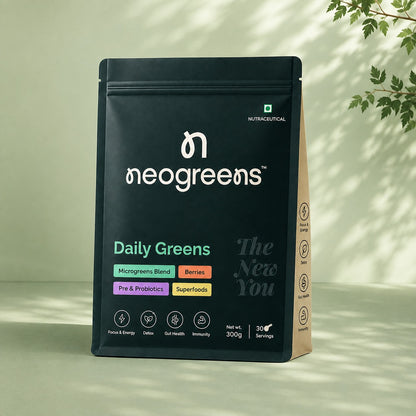 A black stand-up pouch of neogreens Daily Greens dietary supplement, displayed on a light beige surface with soft leaf shadows. The pouch features white branding and colorful accent labels indicating ingredients like microgreens blend, berries, pre & probiotics, and superfoods.