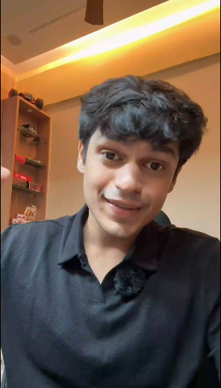 A man wearing a black textured polo shirt, captured in a close-up selfie-style photo inside a home environment with warm lighting and a wooden shelf in the background.